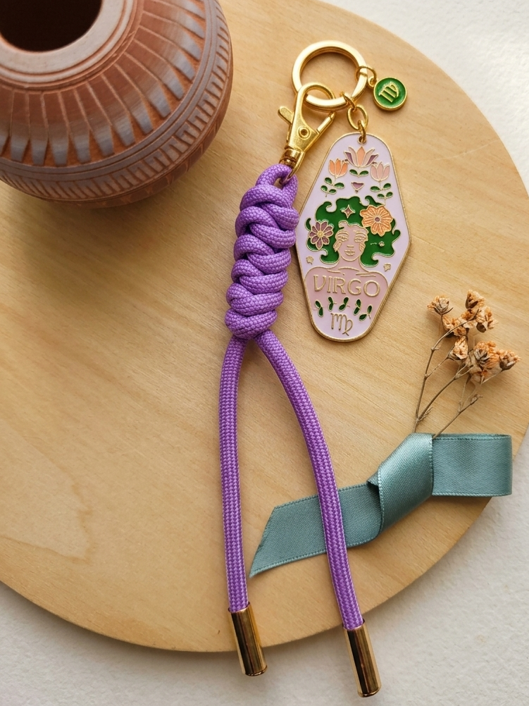 A purple braided cord keychain with a gold clasp and a decorative charm featuring a green turtle and the words 'TIGER TIGER' on a light wood tray, styled with a terracotta vase and dried flowers.