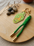 A green and gold keychain with a braided green cord and gold-tone hardware, displayed on a round wooden board with decorative stones and dried stems.